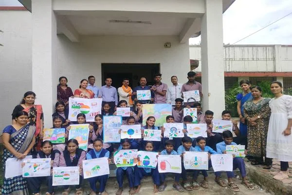 विद्यालय भवन के बाहर अपने बनाए रंगीन चित्र हाथों में लिए बैठे और खड़े विद्यार्थी तथा शिक्षक