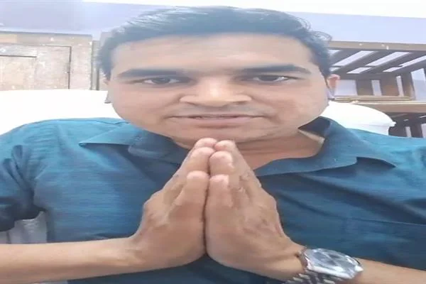 गहरे हरे रंग की शर्ट पहने एक व्यक्ति कैमरे के सामने हाथ जोड़कर बैठा है