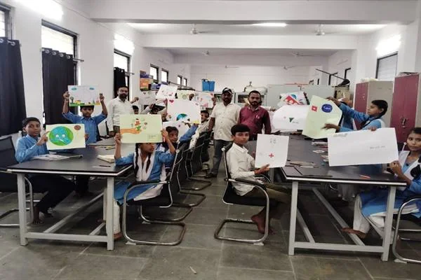 कक्षा में मेज-कुर्सियों पर बैठे विद्यार्थी अपने बनाए चित्र हाथों में दिखा रहे हैं और पीछे शिक्षक खड़े हैं