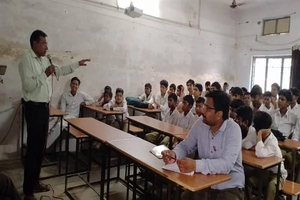 कक्षा में माइक्रोफोन लेकर विद्यार्थियों को संबोधित करते स्वास्थ्यकर्मी और सामने बैठे छात्र