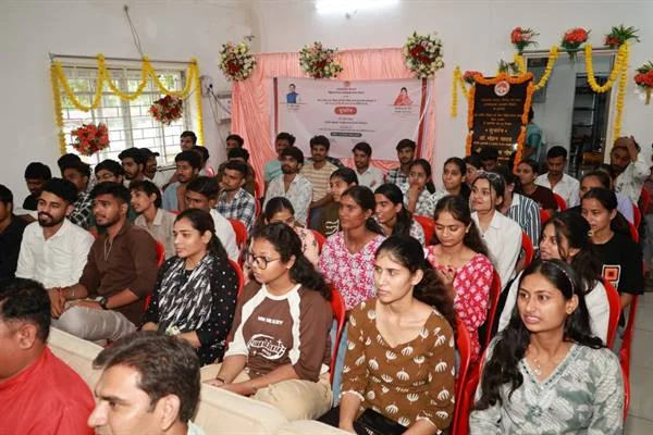 एक हॉल में लाल कुर्सियों पर बैठे छात्र-छात्राएं, पीछे फूलों की मालाओं और बैनर से सजा मंच दिखाई दे रहा है।