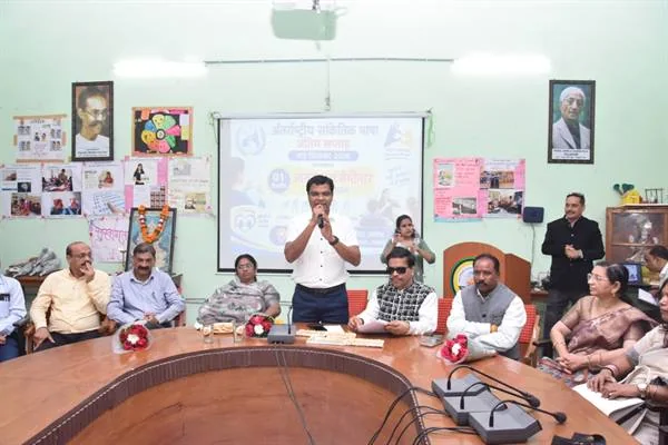 हॉल में गोलाकार मेज के चारों ओर बैठे लोग, बीच में खड़े एक व्यक्ति माइक पर बोलते हुए, पीछे स्क्रीन पर सांकेतिक भाषा से जुड़े कार्यक्रम का बैनर और दीवारों पर पोस्टर
