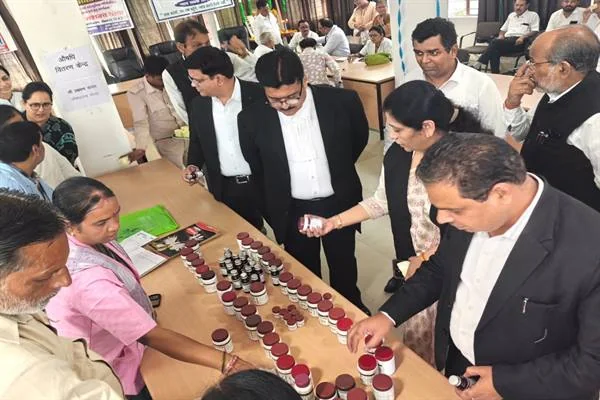 एक हॉल में मेज पर कतार से रखी आयुर्वेदिक दवाइयों की शीशियां, जिनके चारों ओर काले कोट पहने अधिवक्ता और अन्य लोग खड़े होकर दवा ले रहे हैं; पीछे दीवार पर 'औषधि वितरण केंद्र' लिखा कागज लगा है।