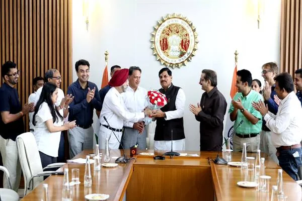 मुख्यमंत्री निवास के बैठक कक्ष में सफेद कुर्ते और काली जैकेट में खड़े मुख्यमंत्री डॉ. मोहन यादव को पगड़ी पहने एक व्यक्ति लाल-सफेद फूलों का गुलदस्ता भेंट कर रहा है; आसपास खड़े कई लोग तालियां बजा रहे हैं और सामने लकड़ी की लंबी बैठक मेज रखी है।